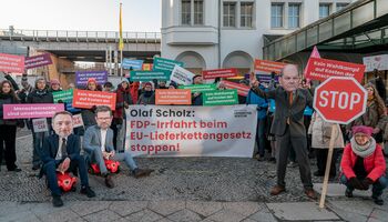 Kundgebung der Initiative Lieferkettengesetz. Im Vordergrund haben sich Personen mit Pappmasken als Christian Lindner, Marco Buschmann und Olaf Scholz verkleidet. Auf einem Transpi steht: Olaf Scholz: FDP-Irrfahrt beim EU-Lieferkettengesetz stoppen!