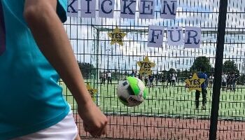 Eine Person schießt einen Fußball gegen einen Zaun auf dem Sportplatz. Am Zaun hängen Zettel mit der Aufschrift Kicken für und Sterne der Initiative Lieferkettengesetz mit der Aufschrift Yes EU can