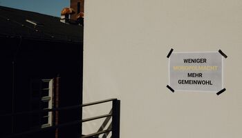 Beschrifteter Zettel auf Hauswand: Weniger Monopolmacht mehr Gemeinwohl