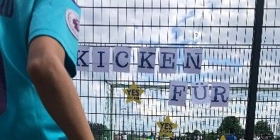 Eine Person schießt einen Fußball gegen einen Zaun auf dem Sportplatz. Am Zaun hängen Zettel mit der Aufschrift Kicken für und Sterne der Initiative Lieferkettengesetz mit der Aufschrift Yes EU can